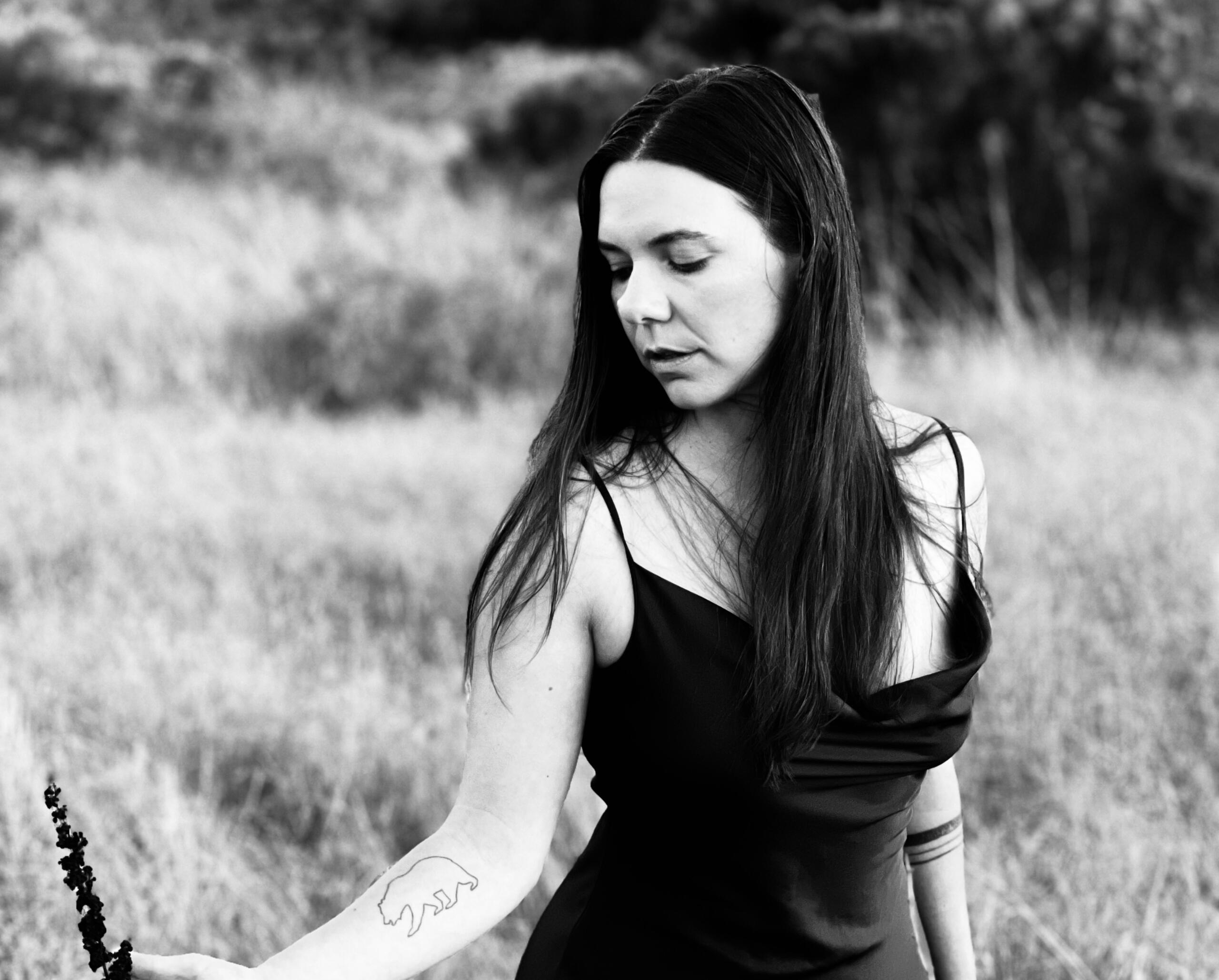 Alex, long-haired with tattoos and a silk dress looking down in a grassy meadow. Black and white